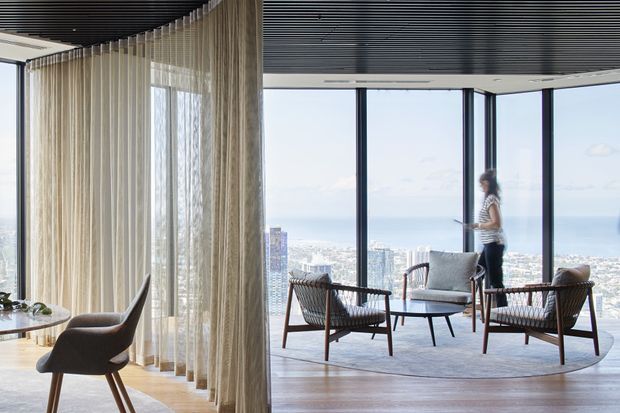 A refined high-rise living room with expansive floor-to-ceiling glazing overlooking a coastal urban skyline and ocean. The room features a curved wall of sheer beige curtains, dark linear ceiling detailing, pale timber flooring, and a contemporary seating arrangement with woven lounge chairs, a sofa, and a round coffee table.