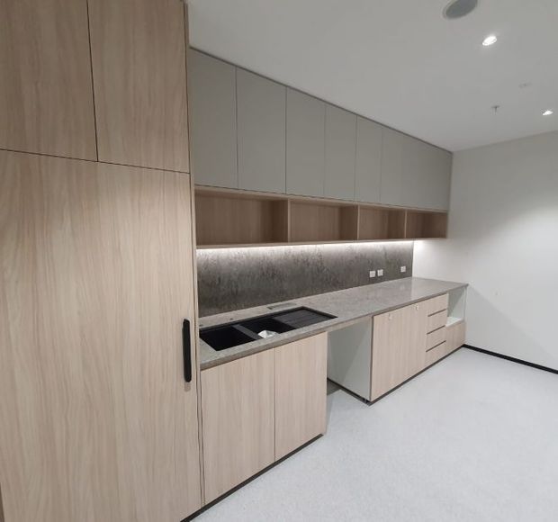 A compact contemporary kitchen or kitchenette with pale timber-grain cabinetry, integrated upper storage, open timber shelving, a grey stone-look benchtop, dark undermount sink, and a textured dark grey splashback. Under-cabinet lighting illuminates the work surface, while the white ceiling and floor create a clean, minimal appearance.