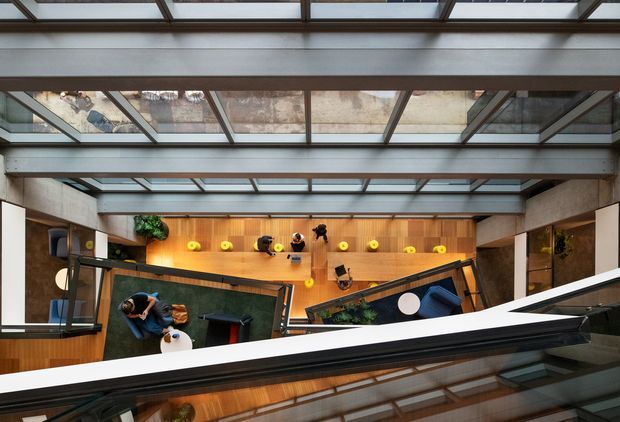 Top-down architectural interior view of a contemporary multi-level living space beneath a large glazed roof or skylight. Exposed grey structural beams frame timber-lined rooms, with dark seating areas, built-in joinery, plants, and warm integrated lighting visible below. A reflective glass or balustrade edge cuts across the foreground.