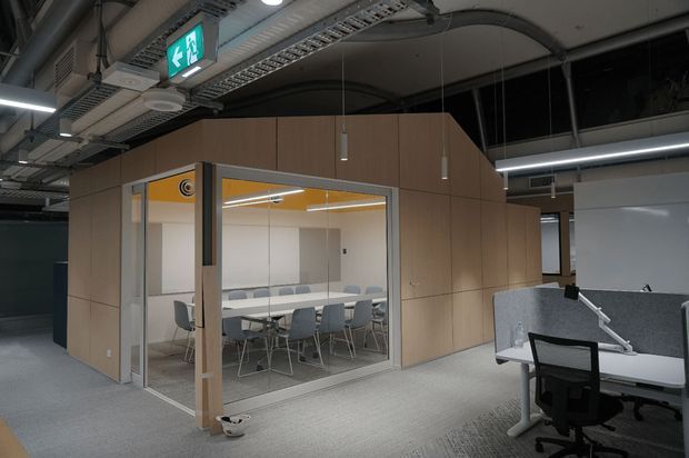 A contemporary commercial office interior with an exposed services ceiling, carpeted floor, open-plan workstations, and a glass-enclosed meeting room clad in light timber panels. The meeting room has a long conference table, multiple chairs, and warm yellow ceiling accents, while adjacent workstations are separated by grey acoustic screens.
