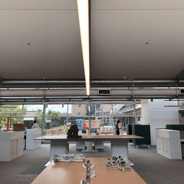 Open-plan commercial showroom or workspace with display counters and cabinetry arranged around a central timber-toned worktable. The space has exposed black ductwork, suspended linear lighting, a dark acoustic ceiling, large glazed façades, and visible shelving or construction displays beyond.