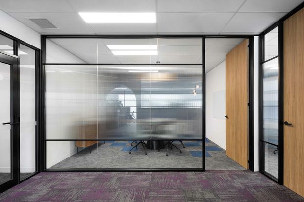 Contemporary commercial office interior viewed from a corridor, featuring black-framed glazed meeting-room partitions with translucent lower panels, timber veneer doors, suspended acoustic ceiling tiles, recessed fluorescent lighting, and patterned grey-purple carpet. Office desks and chairs are visible through the glazing.