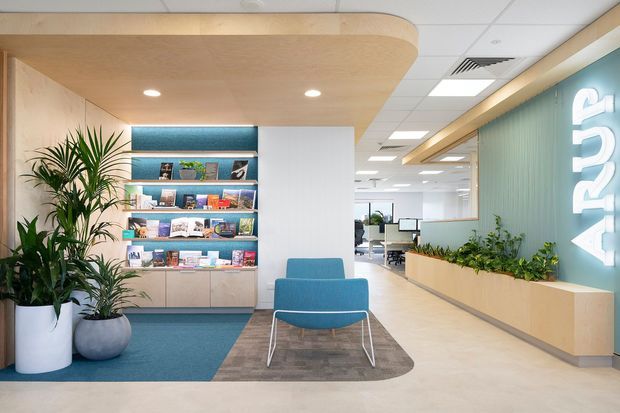 A bright contemporary commercial reception or waiting area with a teal upholstered lounge chair, built-in illuminated display shelving, indoor plants, a curved timber-toned ceiling bulkhead, and a long integrated planter beneath a ribbed teal feature wall with illuminated lettering. An open-plan office area is visible beyond.