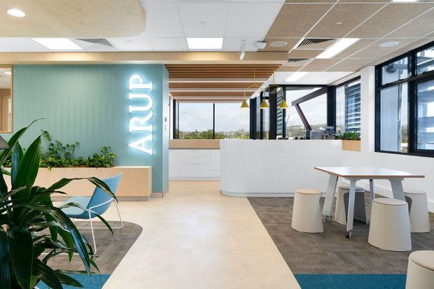 Bright contemporary commercial office reception and breakout area with a curved white reception desk, illuminated ARUP signage, pale timber and teal feature wall, timber ceiling battens, large black-framed windows, indoor planting, and casual dining tables with stools. The space has a clean, open-plan workplace fit-out with views across an urban setting.