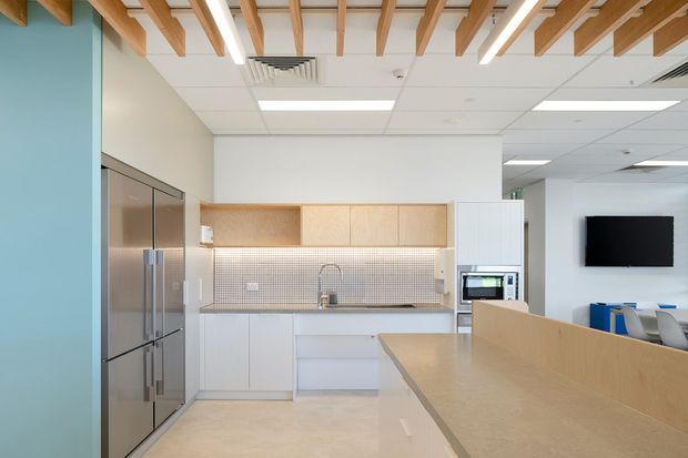 Bright contemporary commercial kitchenette or staff breakout area with white cabinetry, a stainless-steel refrigerator, pale timber open shelving, a small square-tile backsplash, and a long pale timber counter. The space has a white tiled acoustic ceiling with exposed decorative timber beams, linear lighting, and a turquoise feature wall. A microwave, sink, wall-mounted television, and partial blue seating are visible.