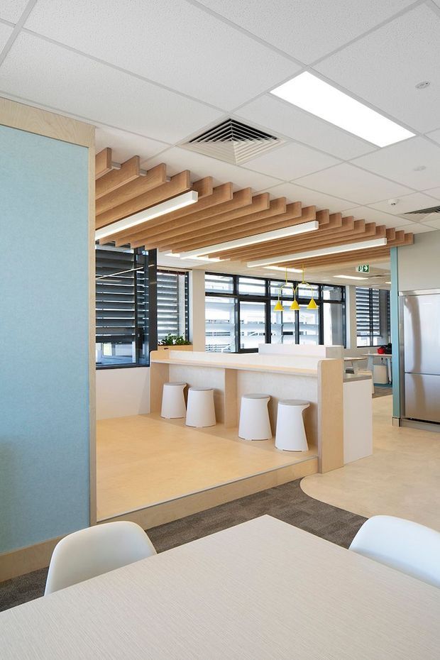 Bright contemporary commercial office breakout and kitchenette area with pale timber finishes, a raised booth-style counter with white stools, communal tables, windows with horizontal blinds, pendant lights, and a striking suspended timber batten ceiling feature beneath a suspended grid ceiling.