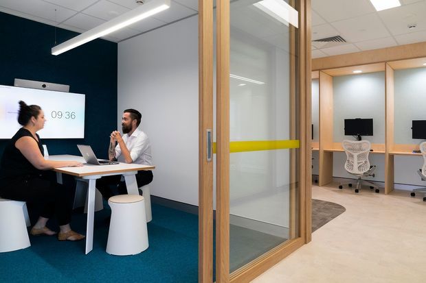 A contemporary commercial office interior showing a collaborative meeting area in the foreground and glazed private workstations beyond. The space features timber-framed glass partitions with a yellow safety stripe, dark teal carpet, white meeting furniture, pale flooring, built-in timber workstation surrounds, ergonomic office chairs, wall-mounted screens, and linear LED lighting in a suspended grid ceiling.