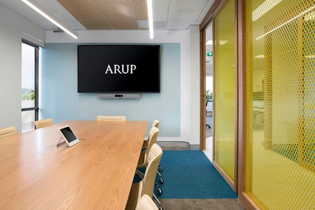 Bright contemporary meeting room with a large light timber conference table, wheeled upholstered chairs, wall-mounted display, linear LED lighting, blue carpet, and a distinctive yellow perforated screen enclosing a glazed office or circulation area. Full-height glazing and clean built-in finishes create a modern commercial workplace setting.