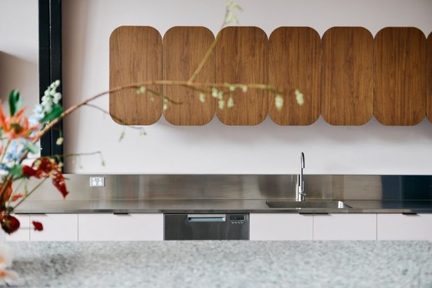 Contemporary kitchen with white flat-panel cabinetry, stainless steel benchtop and splashback, integrated dishwasher, and a distinctive wall-mounted timber feature made from rounded vertical panels. A stone island or benchtop occupies the foreground, with floral branches partially visible at the left.