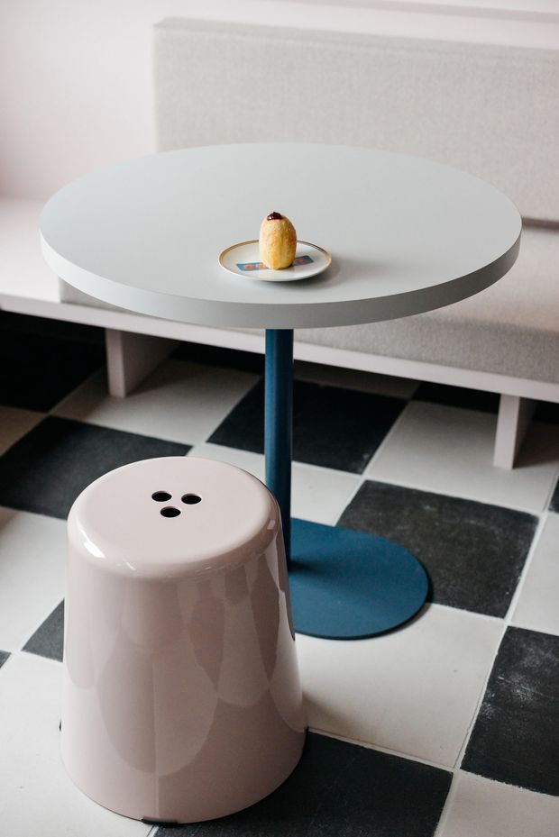 A close-up interior scene featuring a small round white side table with a dark blue central pedestal, a pale pink glossy stool, and a matching blue circular floor base. The furniture sits on a bold black-and-white checkerboard tile floor, with a light upholstered bench or sofa in the background. A small plate and pastry provide a subtle tabletop detail. The composition has a playful, graphic, retro-modern feel.