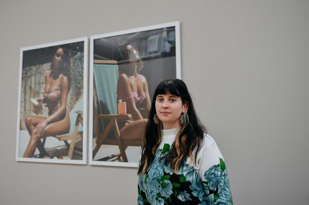 A portrait-oriented photograph of a woman standing indoors in front of two large framed photographic artworks mounted on a plain grey wall. The scene resembles a contemporary gallery or exhibition space, with muted architectural finishes and colorful floral clothing providing the main visual accent.