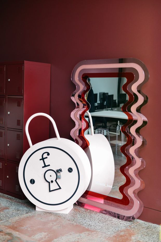 A bold red-toned interior featuring dark red metal lockers, a large sculptural wavy-edged pink and red mirror, and a prominent white safe-shaped decorative object in the foreground. The mirror reflects a workspace or office area with desks and chairs. The space has a playful, highly stylised contemporary fit-out with patterned flooring and strong colour contrast.