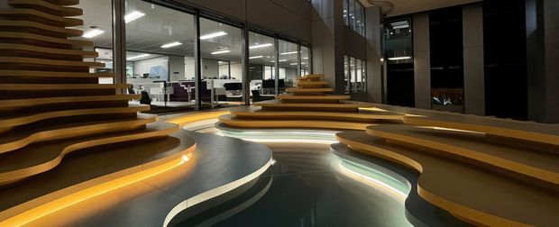 A dramatic contemporary commercial interior featuring sculptural, tiered timber seating and sweeping curved platforms arranged around a glossy dark floor. Warm integrated LED lighting glows beneath the seating edges, while full-height black-framed glazing reveals office workspaces beyond. The space has a refined, futuristic and highly designed appearance with strong flowing forms and layered horizontal lines.