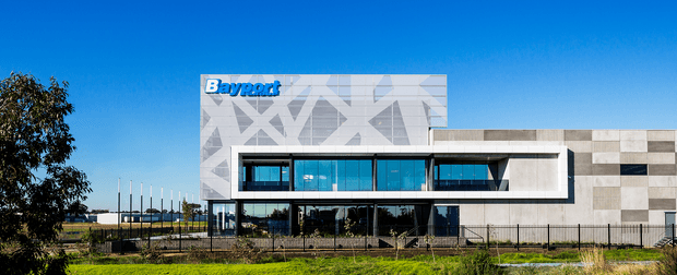 Exterior view of a contemporary commercial building with a bold geometric patterned upper facade, extensive blue-tinted glazing, and a contrasting white rectangular projecting frame. The right wing uses a grid of light and dark rectangular cladding panels. The Bayport sign is visible on the upper facade, with landscaped grounds, fencing, and a clear blue sky.