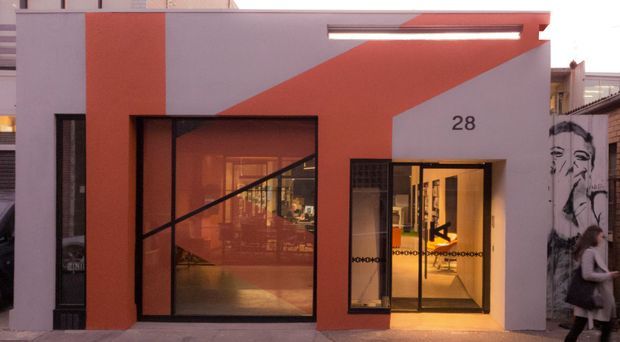 Street-facing contemporary commercial premises with a bold geometric red-orange and grey facade. The building has large black-framed glazed openings, a recessed illuminated entry marked with the number 28, and warm interior lighting visible through the glass. A large monochrome mural is present on the adjacent wall.