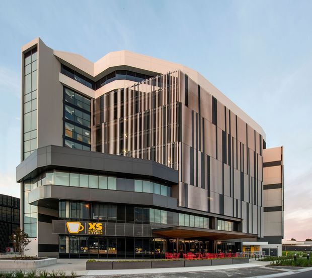 Exterior view of a contemporary multi-storey commercial building with a prominent glazed corner tower, dark framed windows, projecting upper volumes, and a tall vertical fin screen across the main facade. The ground floor contains retail or hospitality frontage with outdoor seating and signage, while paved landscaping surrounds the building.