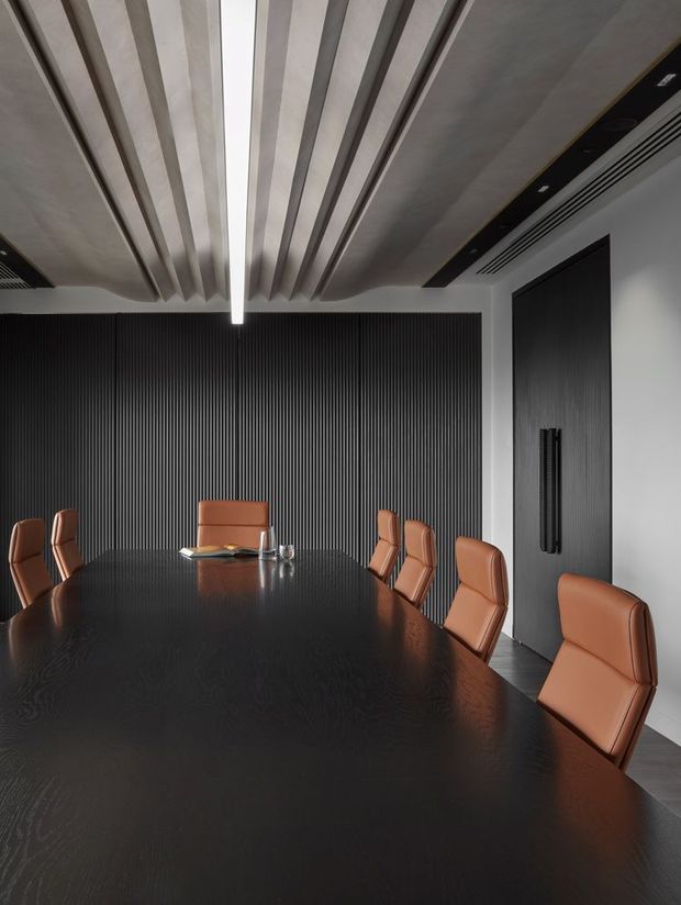 A sophisticated contemporary boardroom with a long dark timber conference table surrounded by tan leather upholstered chairs. The room features a dramatic black vertical fluted feature wall, a dark timber door with elongated handles, white walls, and a sculptural ceiling with parallel recessed beams and a linear illuminated strip. Integrated black ceiling services and air-conditioning contribute to the refined corporate interior.