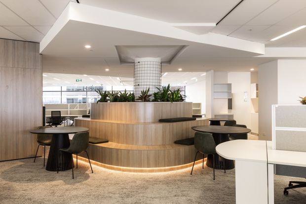 Bright contemporary commercial interior featuring a reception or collaborative lounge area with a curved timber-clad built-in planter and banquette, café-style tables and chairs, carpet flooring, open shelving, and a tiled structural column. Suspended angular white ceiling feature and extensive glazing create an airy, polished workplace environment.
