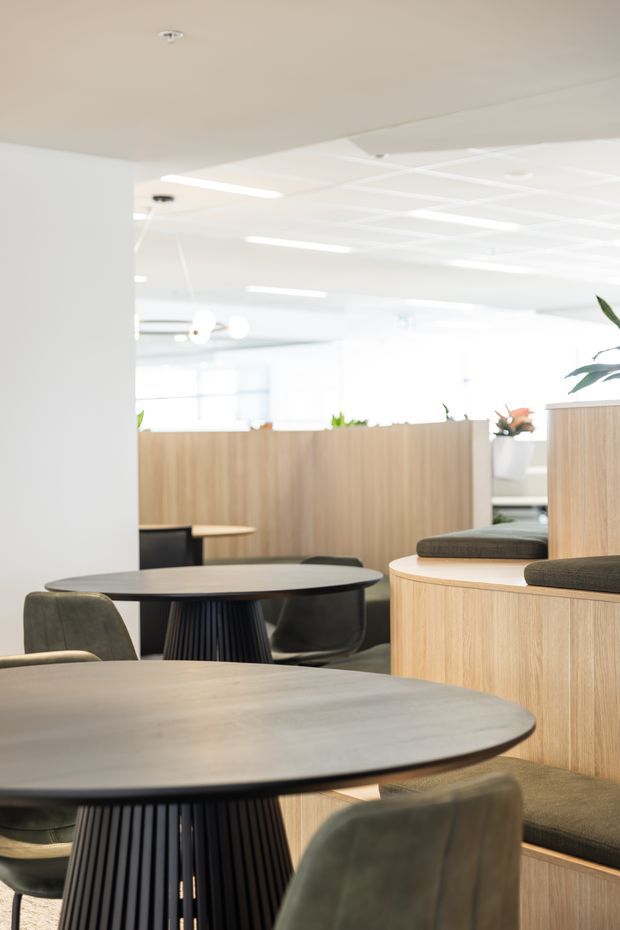 A bright contemporary commercial interior featuring communal dining and work tables with dark round tops and sculptural black pedestal bases. Built-in light-oak partition walls and curved banquette seating define the open-plan space, with soft olive upholstery, indoor plants, and a white suspended ceiling with linear lighting.