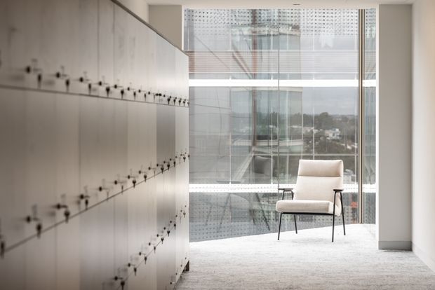 A minimalist office or commercial interior with a long wall of pale built-in cabinetry or storage panels on the left, a large floor-to-ceiling glazed window at the end, and a single light upholstered lounge chair on a pale carpet. The glazing reveals an urban building and landscape beyond, while the room itself has clean white architectural surfaces and a restrained, contemporary appearance.