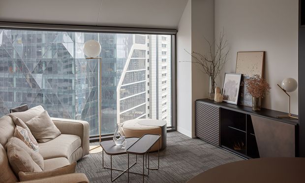 A refined contemporary living room in a high-rise apartment, featuring floor-to-ceiling urban glazing, a neutral upholstered sofa and ottoman, nested coffee tables, a sculptural floor lamp, and a dark textured sideboard styled with framed artwork and dried branches. The room has a calm, sophisticated palette with strong city views and layered textures.