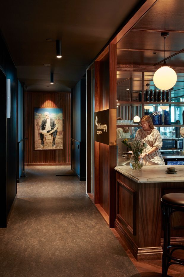 A moody, richly detailed commercial hospitality interior showing a bar or café counter opening onto a dark timber-lined corridor. The space features extensive warm wood panelling and cabinetry, a pale stone or concrete-look benchtop, dark flooring, black-framed glazing, brass-toned details, and spherical pendant lighting. A bartender is arranging flowers behind the counter, with bottles, glassware, and café equipment visible. A framed artwork anchors the corridor at the rear.