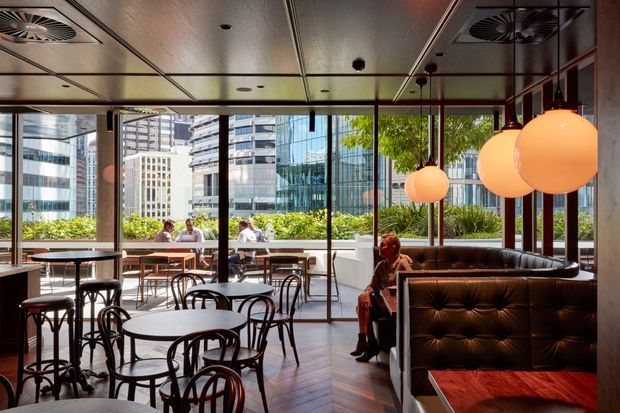 A contemporary urban café or restaurant interior with floor-to-ceiling glazing, outdoor terrace seating, and views of surrounding high-rise buildings. The space features dark timber flooring, black bentwood-style chairs and round tables, tufted dark banquette seating, warm pendant globes, exposed dark framing, and a reflective metal ceiling with integrated ventilation. Bright daylight contrasts with the warm, intimate interior.