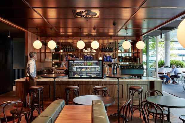 A warmly lit contemporary café interior with a timber-lined ceiling and bar counter, pendant globe lights, dark timber detailing, marble or stone benchtops, and café seating. The central service counter includes a coffee machine, display case, shelving, and bottle storage, while large glazed openings connect to an outdoor seating area.