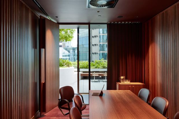 A refined contemporary meeting or dining room clad in rich dark timber wall panelling, with a long timber table and upholstered chairs. Full-height glazing opens to a planted terrace and an urban city setting beyond. The ceiling incorporates recessed lighting and a visible ventilation grille, creating a warm, sophisticated commercial interior.