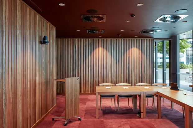 A refined meeting or dining room with full-height vertical timber wall panelling, a deep burgundy ceiling and carpet, and a long timber table surrounded by chairs. A timber lectern stands at the left, while large glazed openings on the right provide views to an urban exterior. Recessed and ceiling-mounted services create a commercial interior atmosphere.
