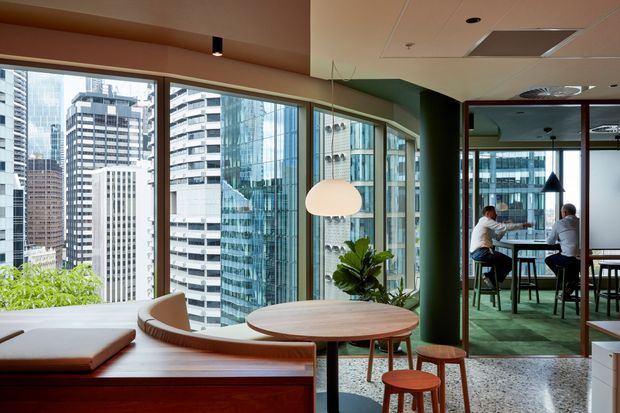 A contemporary office lounge and meeting area with expansive curved glazing overlooking a dense city skyline. The space features built-in timber seating, a round timber table, stools, a sculptural pendant light, green carpet, a dark green structural column, and a glazed meeting room beyond.
