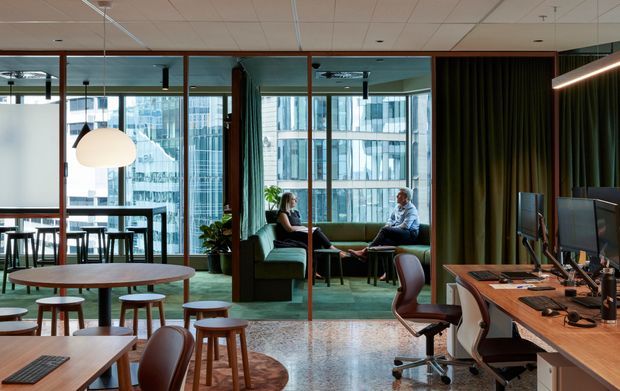 A contemporary commercial office interior with an open lounge and informal meeting area beside a workstation zone. The space features floor-to-ceiling glazing with views of surrounding office buildings, dark green carpet, timber-framed glass partitions, warm wood desks, leather task chairs, pendant lighting, and a dark curtained meeting or presentation area. Two people are seated in the lounge, with a communal table and stools in the foreground.