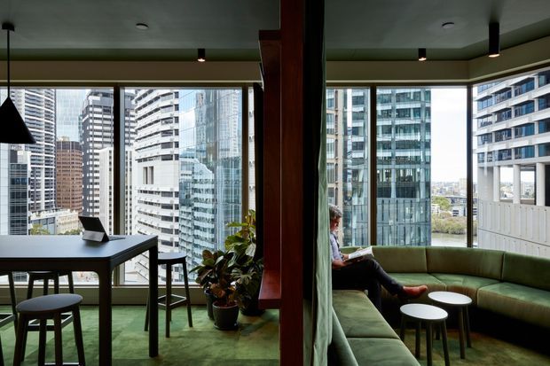 A sophisticated high-rise apartment interior with an open-plan living and dining area framed by extensive corner glazing. The space features a green upholstered sectional sofa, matching green carpet, a dark dining table with stools and chairs, small round coffee tables, indoor plants, dark timber-toned structural or joinery elements, and pendant and recessed lighting. Dense urban towers are visible outside the windows, giving the room a metropolitan outlook.