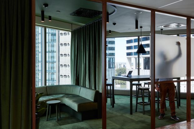 A sophisticated contemporary commercial interior viewed through timber-framed glazed partitions. The space includes a green upholstered curved banquette, dark green carpet and curtains, a high communal table with stools, pendant lighting, and floor-to-ceiling windows overlooking an urban setting. A frosted glass partition with a blurred figure adds privacy while maintaining light.