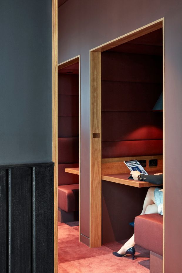 A compact, richly coloured private booth or workspace viewed through a timber-framed opening. The space features deep burgundy upholstered wall panels and seating, red carpeting, dark mauve painted walls, warm timber shelves and desk surfaces, and integrated power outlets. A person is seated at a small desk using a laptop or tablet. A tall mirror reflects the adjacent upholstered booth, emphasizing the enclosed, bespoke joinery and intimate atmosphere.