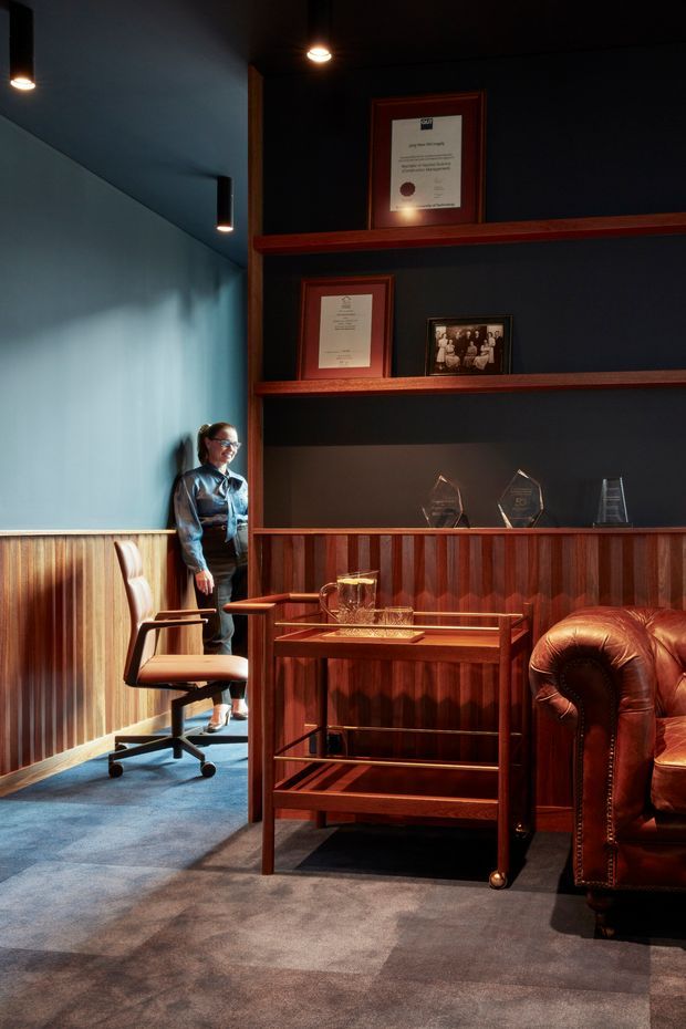 A moody, richly detailed interior with dark navy walls, warm timber wall panelling and built-in display shelving, showcasing framed certificates, photographs and awards. A vintage leather Chesterfield-style armchair and timber drinks trolley sit on a patterned grey carpet, while a small adjoining workspace contains a leather office chair. Warm recessed and pendant lighting creates a refined, intimate atmosphere.