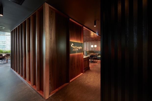 A moody contemporary commercial interior featuring a timber-lined partition and reception or entry zone. Rich dark wood vertical battens frame a partially enclosed dining or hospitality area, with warm pendant lighting, polished dark flooring, and a branded sign on the timber wall. Glazing at the left opens toward a bright seating area, creating strong contrast between the shadowed corridor and illuminated interior.