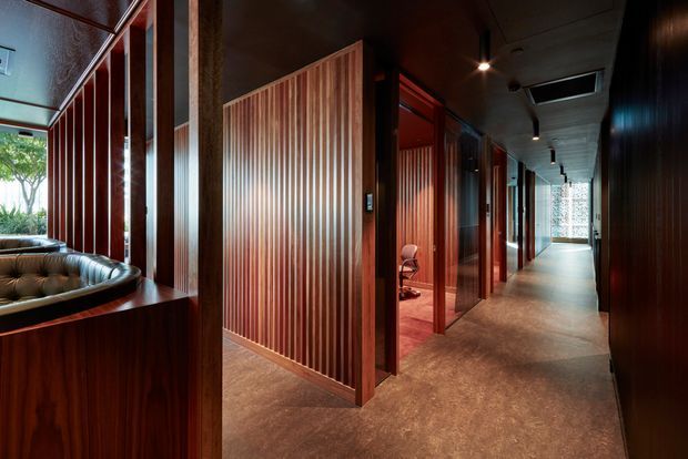 A dark, richly timber-lined interior corridor with a series of private rooms or offices opening along one side. Vertical timber slat partitions, deep reddish-brown wood wall panelling, a polished concrete or stone-look floor, recessed ceiling lighting, and glimpses of upholstered seating create a refined commercial or hospitality atmosphere. Daylight enters through glazing at the far end and from the left side.