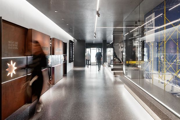A polished contemporary commercial interior, likely a museum, gallery, or branded workplace lobby. The long corridor has a terrazzo-like grey floor, dark concrete ceiling, linear recessed and suspended lighting, and illuminated timber-toned display panels along the left wall. On the right, full-height glazing reveals a graphic blue-and-yellow installation or exhibition display, while several people move through the space.