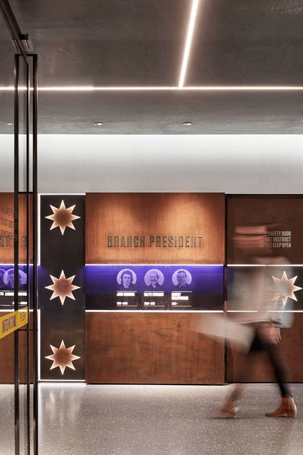 A dramatic commercial interior featuring a polished speckled floor, dark textured ceiling with linear recessed LED lighting, glass doors and partitions, and a wood-paneled wall displaying “BRANCH PRESIDENT” and historical portraits. Starburst wall lights, purple accent illumination, dark metal framing, and reflective surfaces create a refined, museum-like lobby or exhibition corridor.
