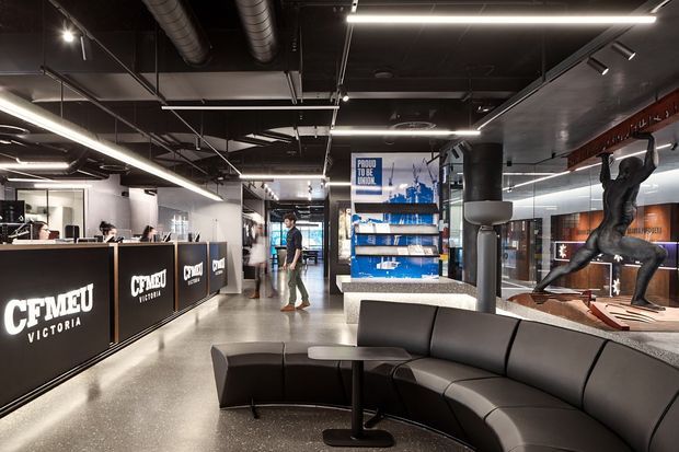 A contemporary commercial reception and waiting area for CFMEU Victoria. The space features a large black reception desk with illuminated branding, polished concrete flooring, exposed black ceilings with visible ductwork, linear and track lighting, a curved black leather sectional sofa, display shelving, and a prominent sculptural figure. Glazed office or meeting spaces are visible in the background.