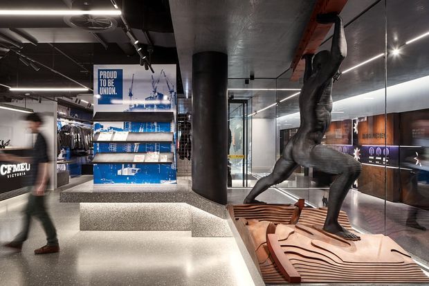A dramatic contemporary commercial interior resembling a sports club or branded visitor centre. The space features dark exposed services and ceilings, linear LED lighting, polished terrazzo-like flooring and stepped seating, glass partitions, illuminated product displays, and a large sculptural athlete figure climbing overhead. Branded merchandise and graphics are visible in the background.