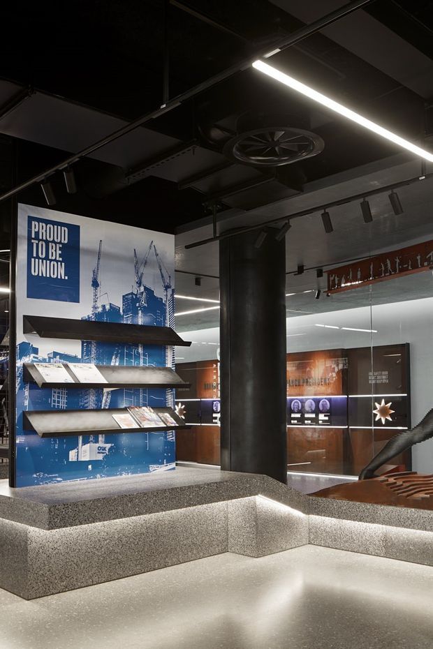 A dark, industrial-style commercial interior resembling a branded visitor centre, showroom, or exhibition space. The foreground features a large stepped terrazzo display platform with integrated linear lighting, supporting a graphic display rack with construction imagery and printed materials. Black exposed ceiling services, ducts, track lighting, a prominent cylindrical column, timber-toned wall panels, illuminated graphics, and sculptural seating create a contemporary industrial fit-out.