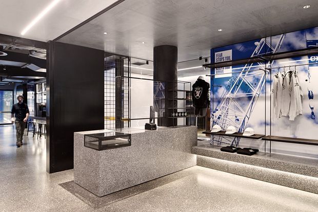 A contemporary commercial retail or showroom interior with a polished terrazzo reception/display counter in the foreground. The space features dark charcoal wall panels and columns, a reflective metallic ceiling, linear recessed lighting, tiled and gridded partitions, and a large blue-and-white branded sports display wall with hanging apparel. Built-in illuminated display plinths and benches define the showroom layout.