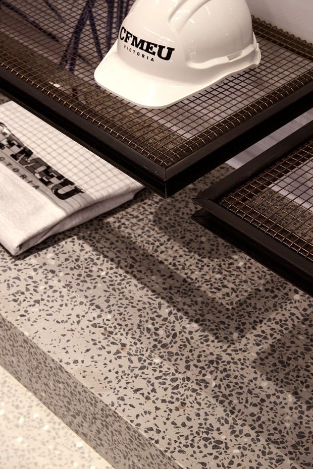 Close-up product and materials photograph showing a white construction hard hat marked “CFMEU Victoria” resting on stacked black metal mesh display trays. A folded printed textile is partially visible at left. The dominant architectural finish is a light terrazzo surface with dense dark grey aggregate, including a thick stepped or slab edge. The image is styled like a construction-material showroom or trade display, with no identifiable room layout visible.