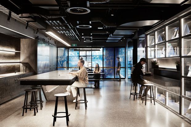 A contemporary commercial interior resembling a collaborative workplace café or staff breakout area. The open-plan space features a long stone island with stools, additional high tables, polished concrete flooring, dark exposed services and ceiling panels, linear and circular pendant lighting, glazed partitions, and illuminated built-in shelving. Several people are using the work and seating areas.