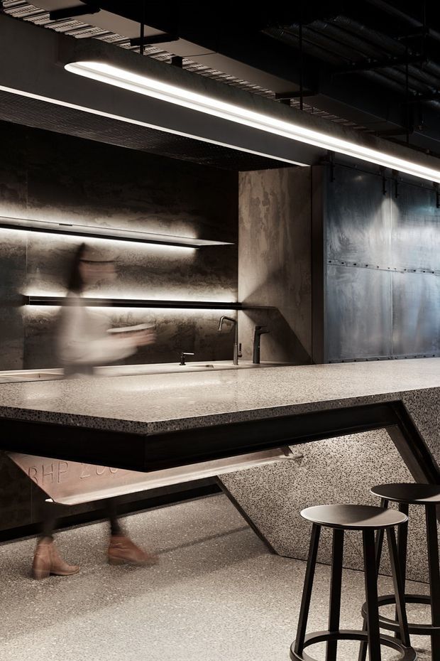 A dark, industrial-style commercial interior centred on a large speckled stone or terrazzo counter with integrated sinks and black angular supports. The space features raw dark walls, exposed ceiling services, linear pendant lighting, metal wall panels, open shelving with concealed lighting, and black bar stools. Blurred people suggest an active hospitality or workplace setting.