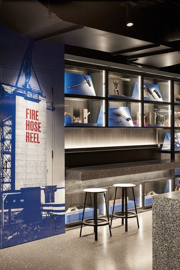 A dark, industrial-style commercial interior with a display wall of illuminated glass cabinets containing tools and equipment. A large blue-and-white graphic door marked “FIRE HOSE REEL” forms a bold feature, while black ceilings, exposed services, metal shelving, terrazzo-like counters, and a pair of simple black stools reinforce the utilitarian workshop or showroom character.