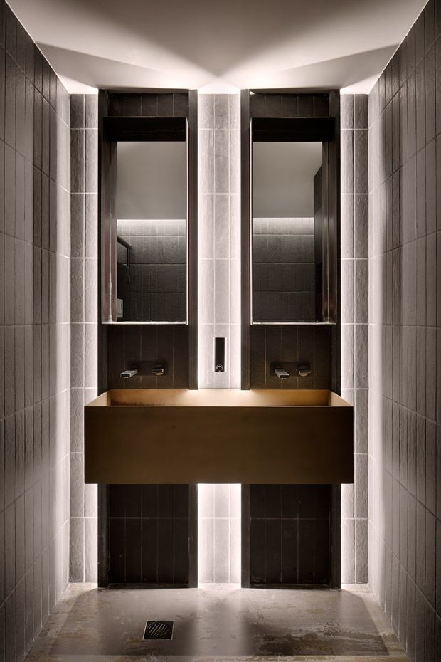 A narrow, symmetrical contemporary bathroom with twin wall-mounted basins, mirrored recessed niches, dark tiled walls, pale vertical tiled columns, and integrated warm strip lighting. The substantial bronze-toned vanity projects across the centre, while a floor drain and wet-room style floor are visible below.