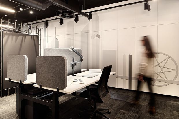 A contemporary open-plan office workspace with shared white desks, black task chairs, acoustic privacy screens, desk lamps, exposed ceiling services, track lighting, and a large monochromatic graphic wall. The space has an industrial-influenced fit-out with dark carpet tiles and black structural elements.